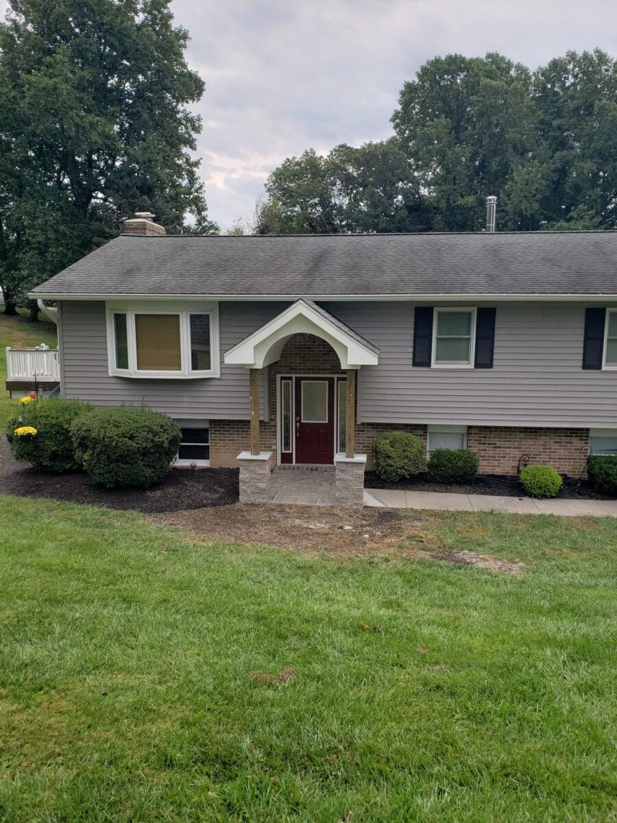 Gray house with white trim, red front door, and a small covered entry surrounded by lawn and trees.