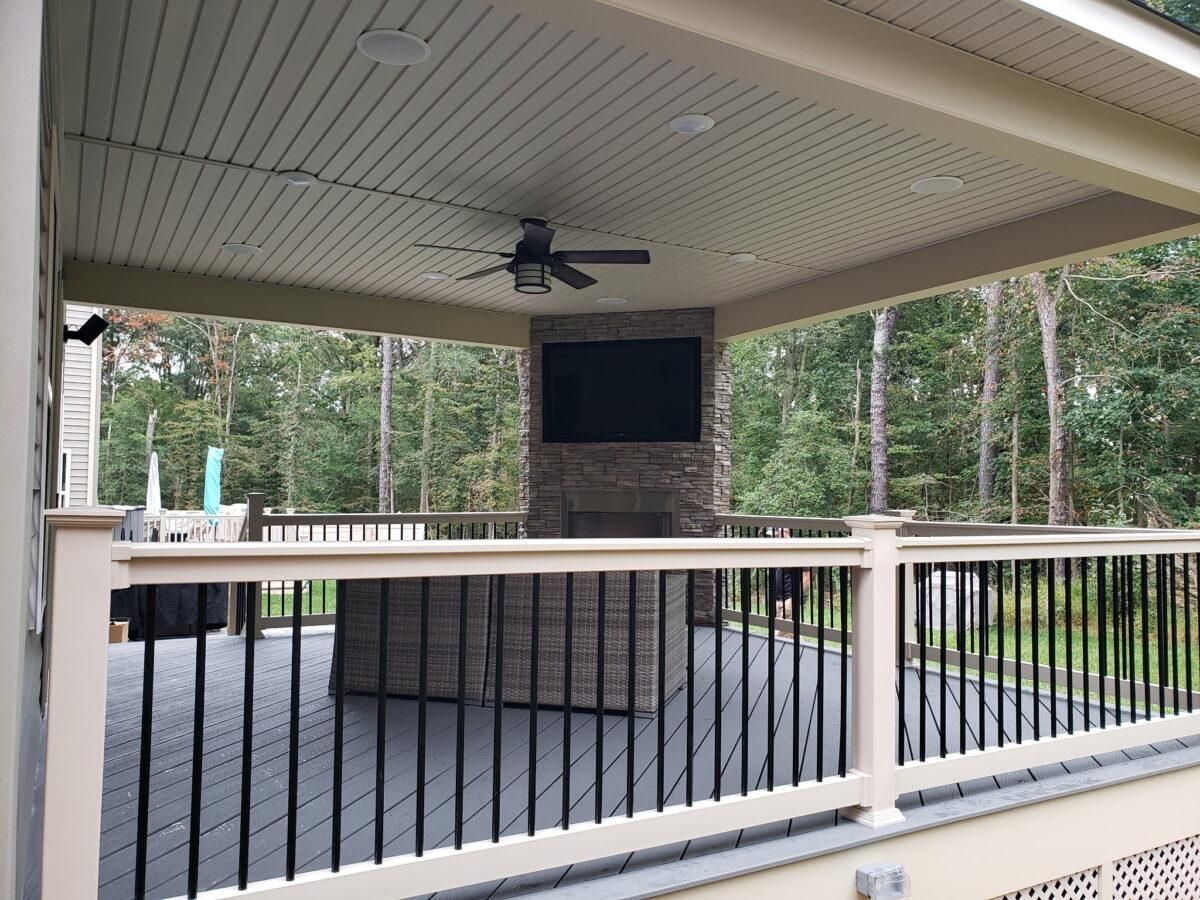 Covered outdoor patio with ceiling fan, TV, fireplace, and railing overlooking trees