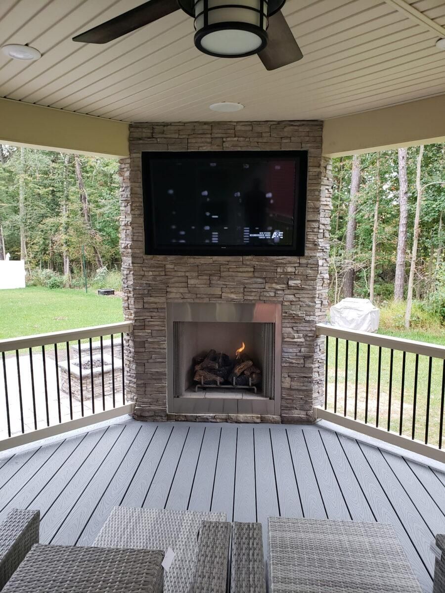 Covered porch with a stone fireplace and mounted TV overlooking a grassy yard and trees.