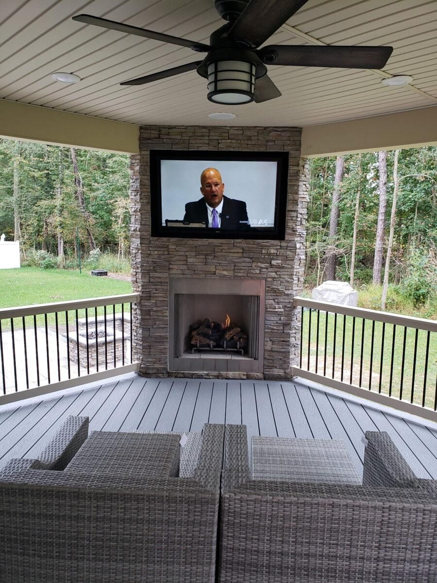 Covered porch with TV above a stone fireplace, wicker seating, and wooded yard beyond