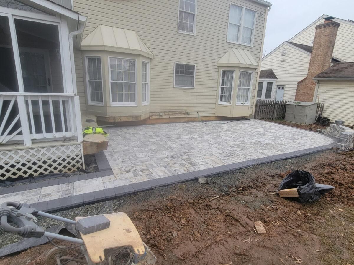 Backyard patio with gray pavers beside a house, bordered by a strip of bare soil under construction