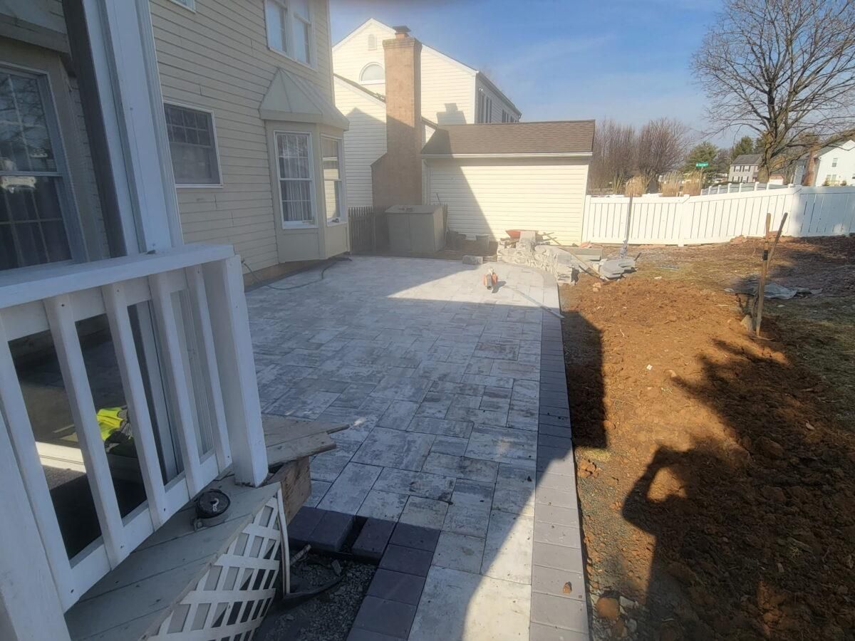 Paver patio beside a house, with a fenced yard and bare trees in winter.