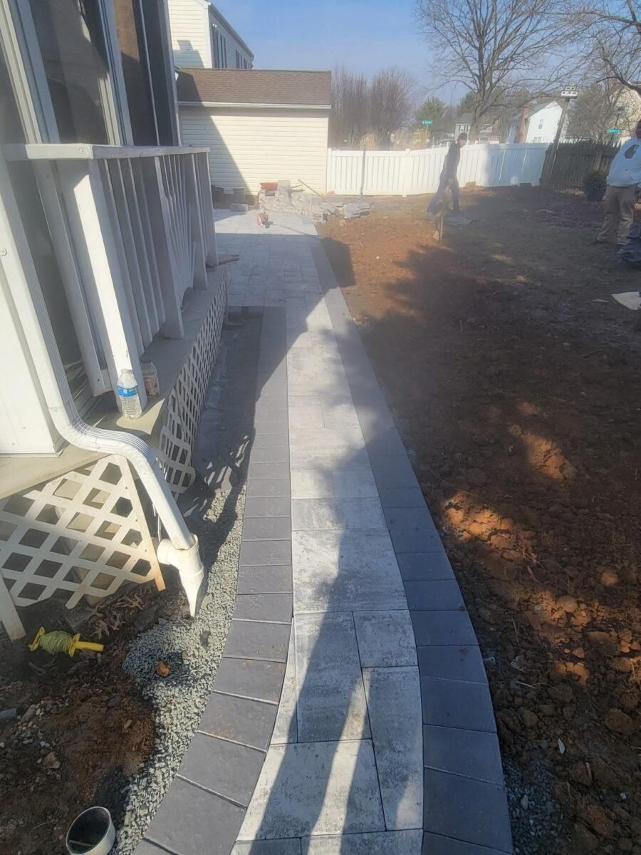 Curved concrete walkway beside a house, bordered by a white fence and bare backyard soil.