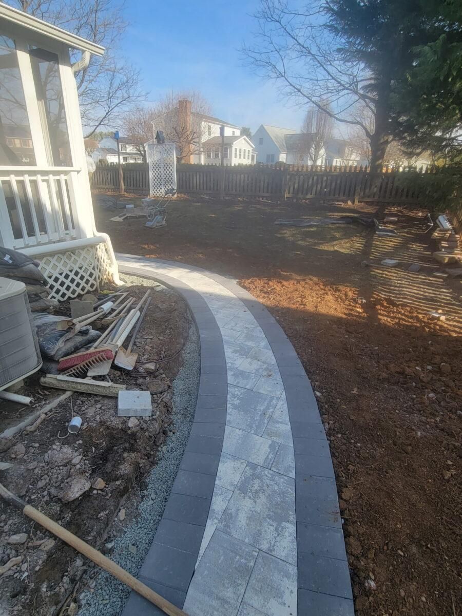 Curved gray stone walkway beside a house, leading through a leaf-covered backyard with a fence and trees.