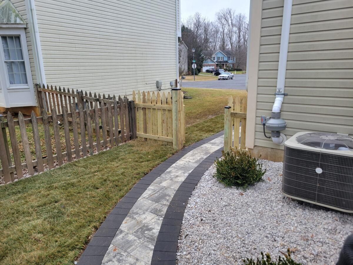 Curved walkway beside a house, with gravel edging, small shrubs, and a wooden fence gate.