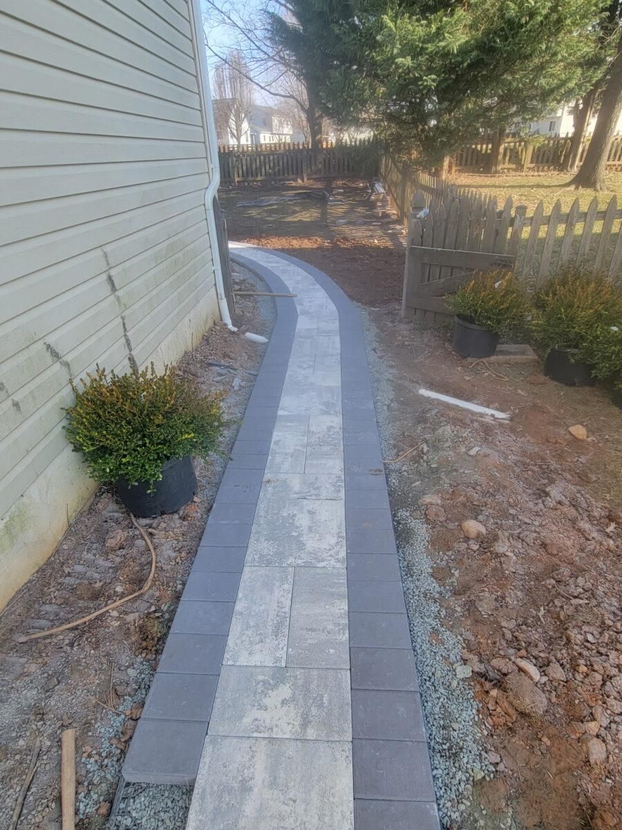 Narrow stone walkway curving beside a house, with a fence and shrubs along the path.