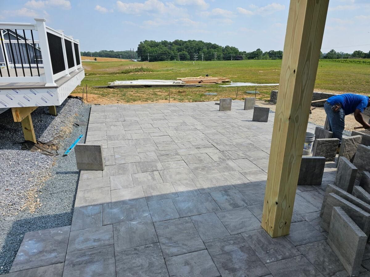 Partially paved patio beside a house, with a worker laying pavers and open grassy land beyond