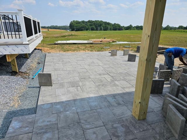 Patio paving stones being installed beside a house, with workers and stacked pavers in a grassy yard.