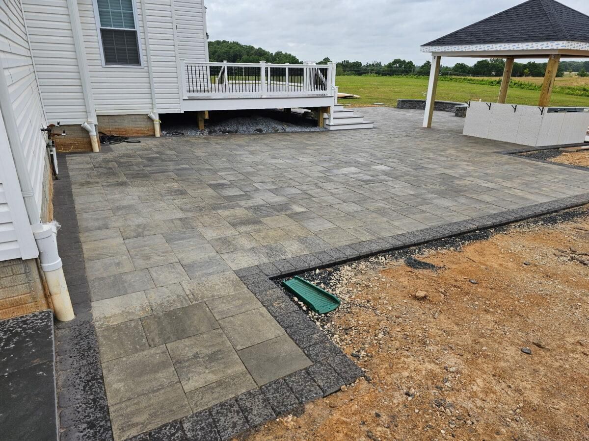 Stone paver patio beside a white house, with a small deck and gravel edging under construction