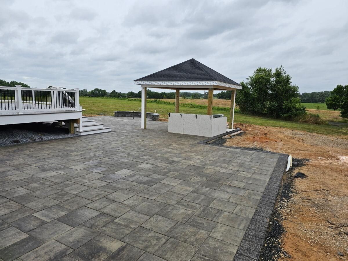 Paved patio with white gazebo and deck overlooking a grassy field under cloudy sky