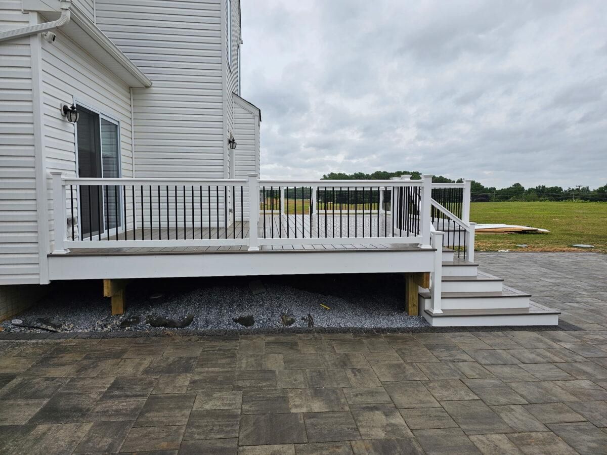 White house deck with railing and steps beside a paved patio, overlooking an open grassy yard.