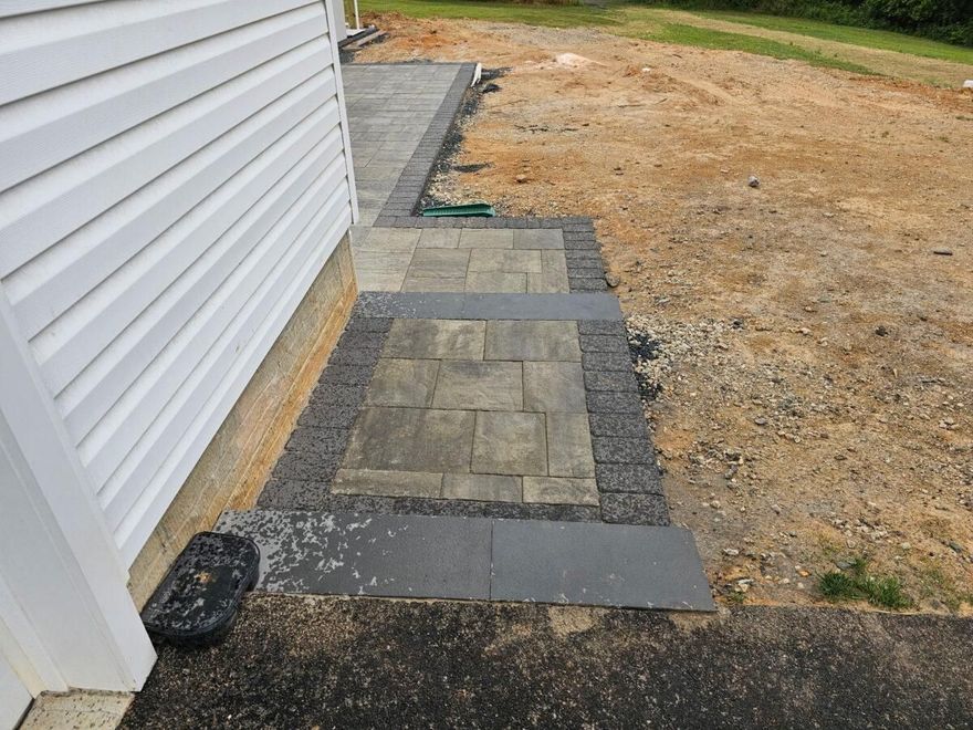 Concrete walkway beside white siding, with dark edging stones leading through a dry yard.