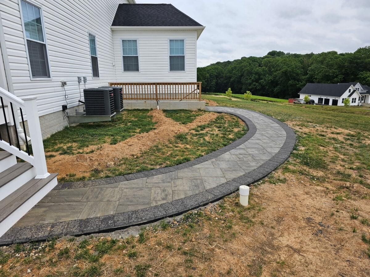 Curved stone walkway beside a white house in a dirt yard, leading toward a deck and distant trees.