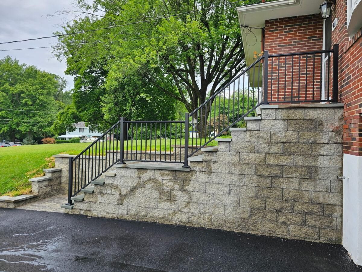 Stone retaining wall with black metal railings and stairs beside a brick porch, next to a wet driveway.