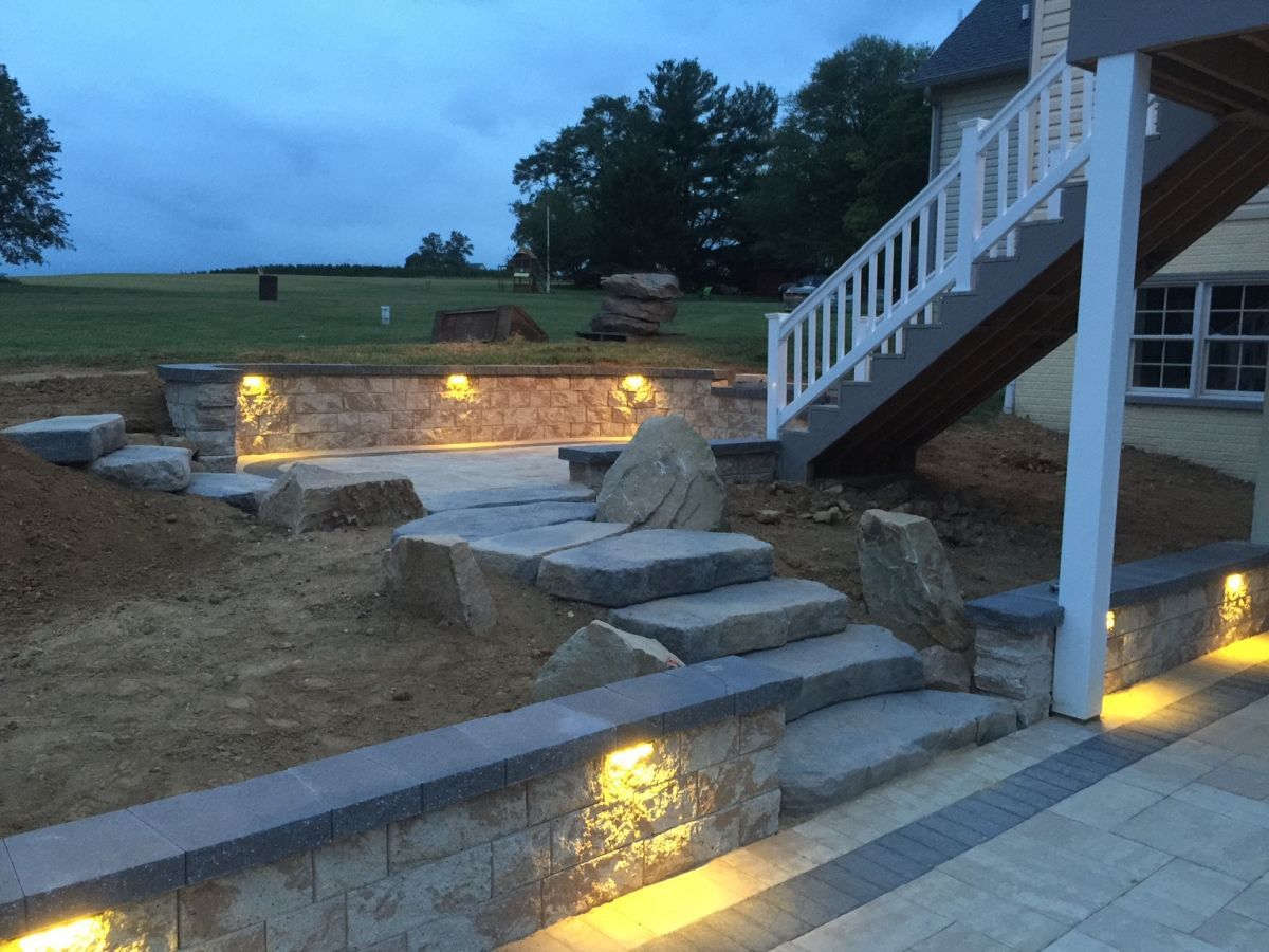Stone patio steps and retaining walls with warm landscape lights beside a white-railed deck at dusk