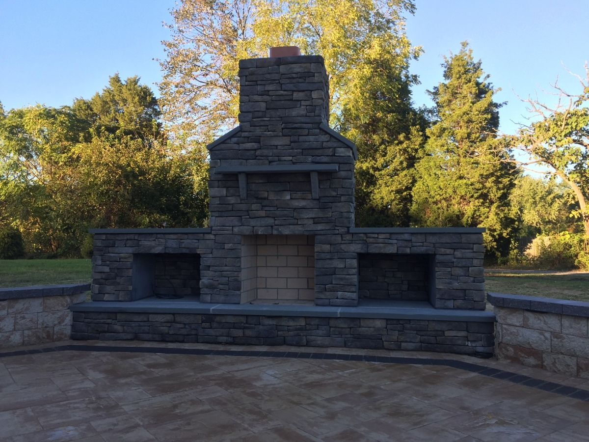 Stone outdoor fireplace with wide chimney and built-in seating in a park, surrounded by trees and sunlight