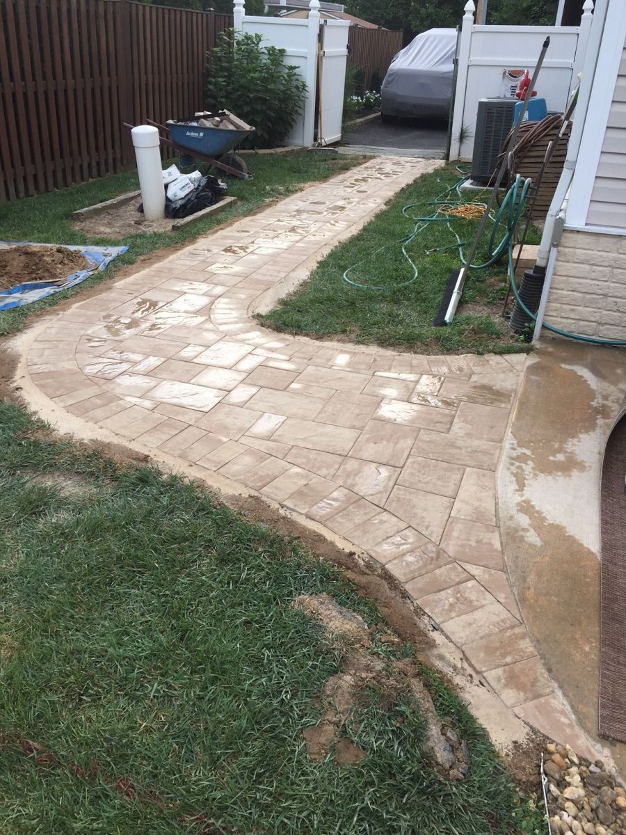 Brick walkway beside a house, bordered by grass, with a wheelbarrow and garden tools in the yard.