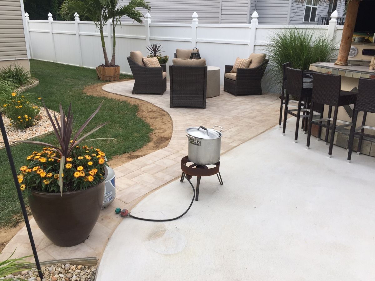 Patio with wicker seating, potted flowers, and a silver pot on a stand connected by a hose on the concrete patio.