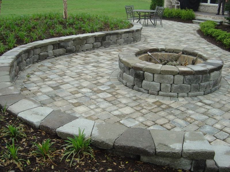 Stone patio with circular fire pit, curved retaining wall, and outdoor table and chairs in a garden