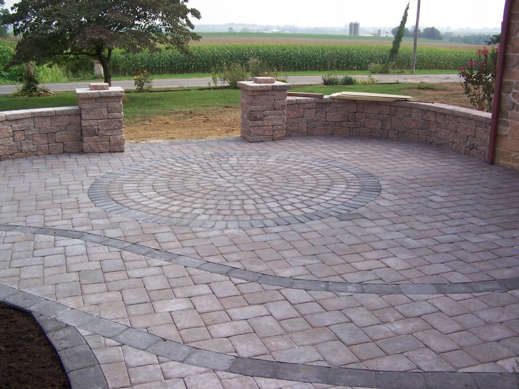 Stone-paved patio with a circular pattern, bordered by low brick walls, overlooking a green field.