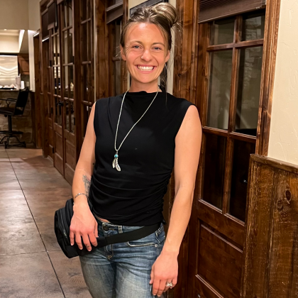 Smiling person in a black sleeveless top and jeans standing in a wood-paneled hallway.