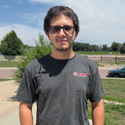 Man in glasses and gray T-shirt standing outdoors on a sunny day near a parking lot and grassy field
