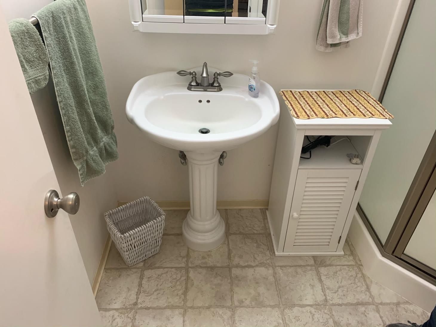 Small bathroom with pedestal sink, towel rack, wicker hamper, and white cabinet with bamboo top.