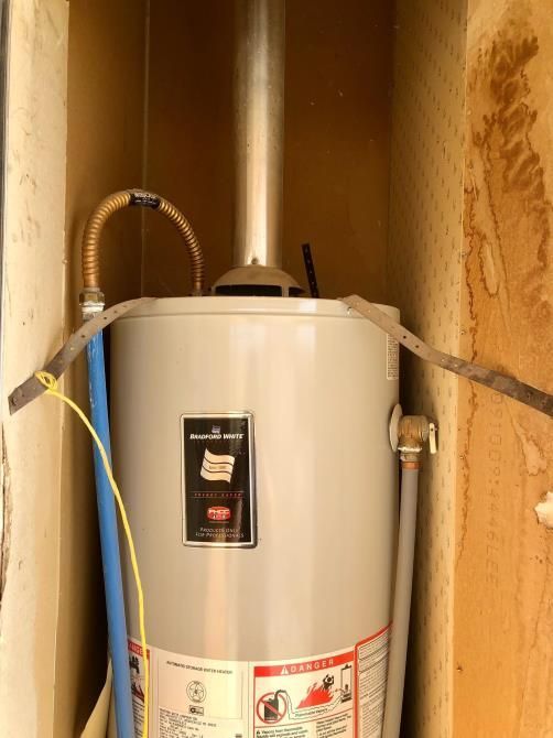 Water heater in a narrow utility closet with exposed pipes and a flexible hose.