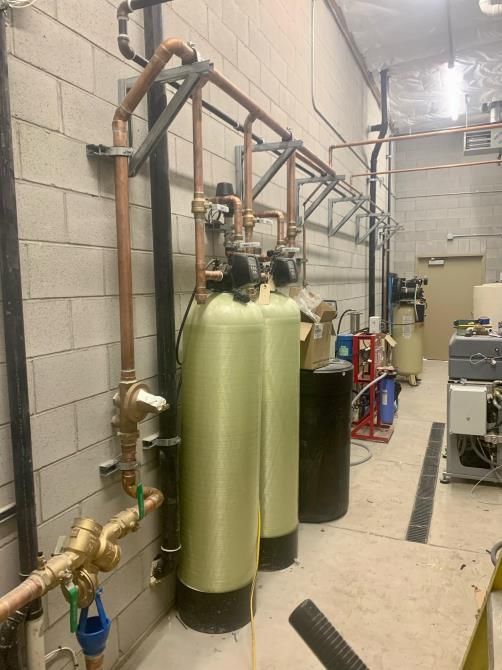 Industrial water treatment tanks and piping in a utility room, with tall green and black cylinders beside cinderblock walls
