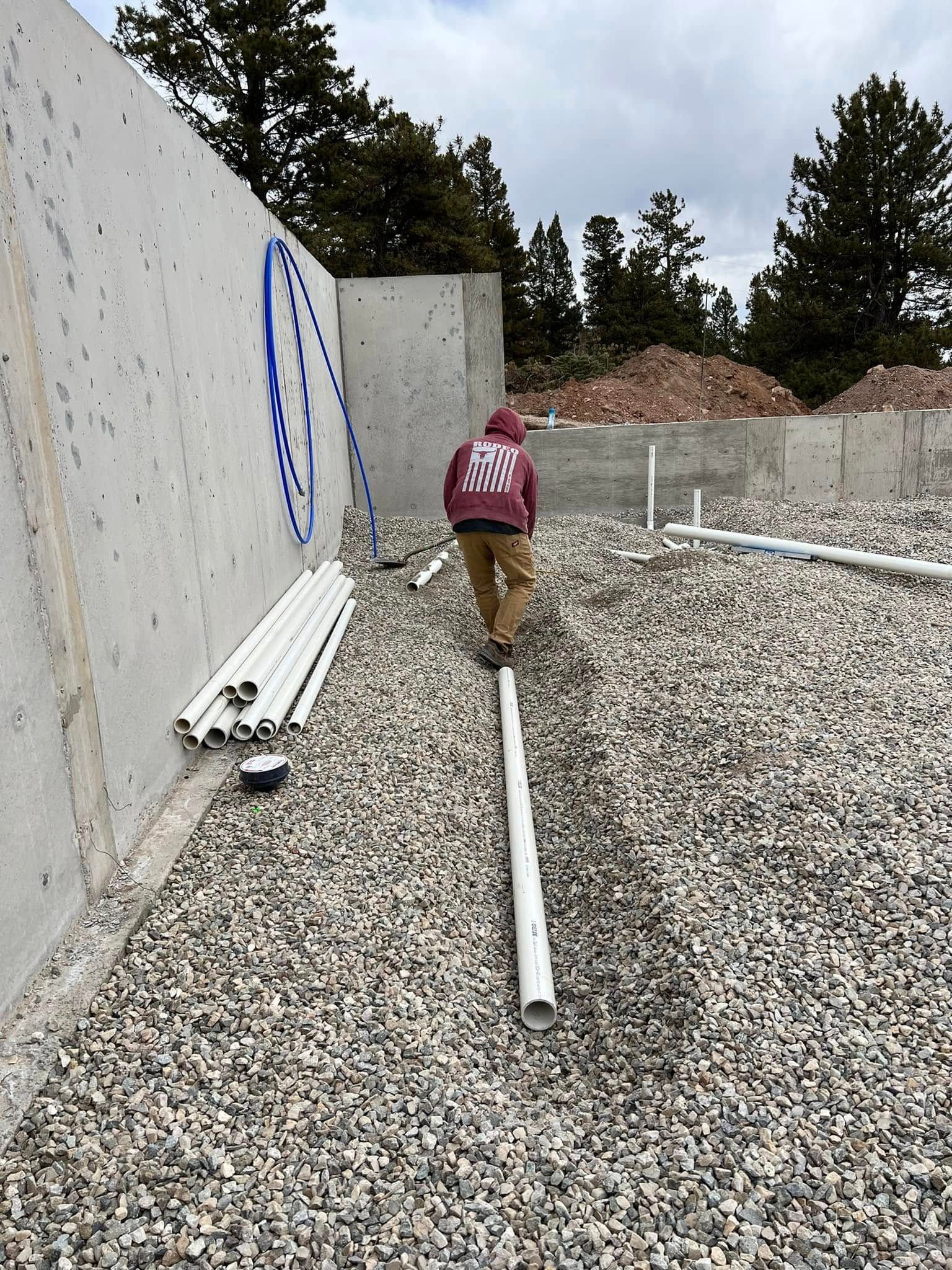 Person walking through a gravel construction site beside concrete walls and scattered pipes.