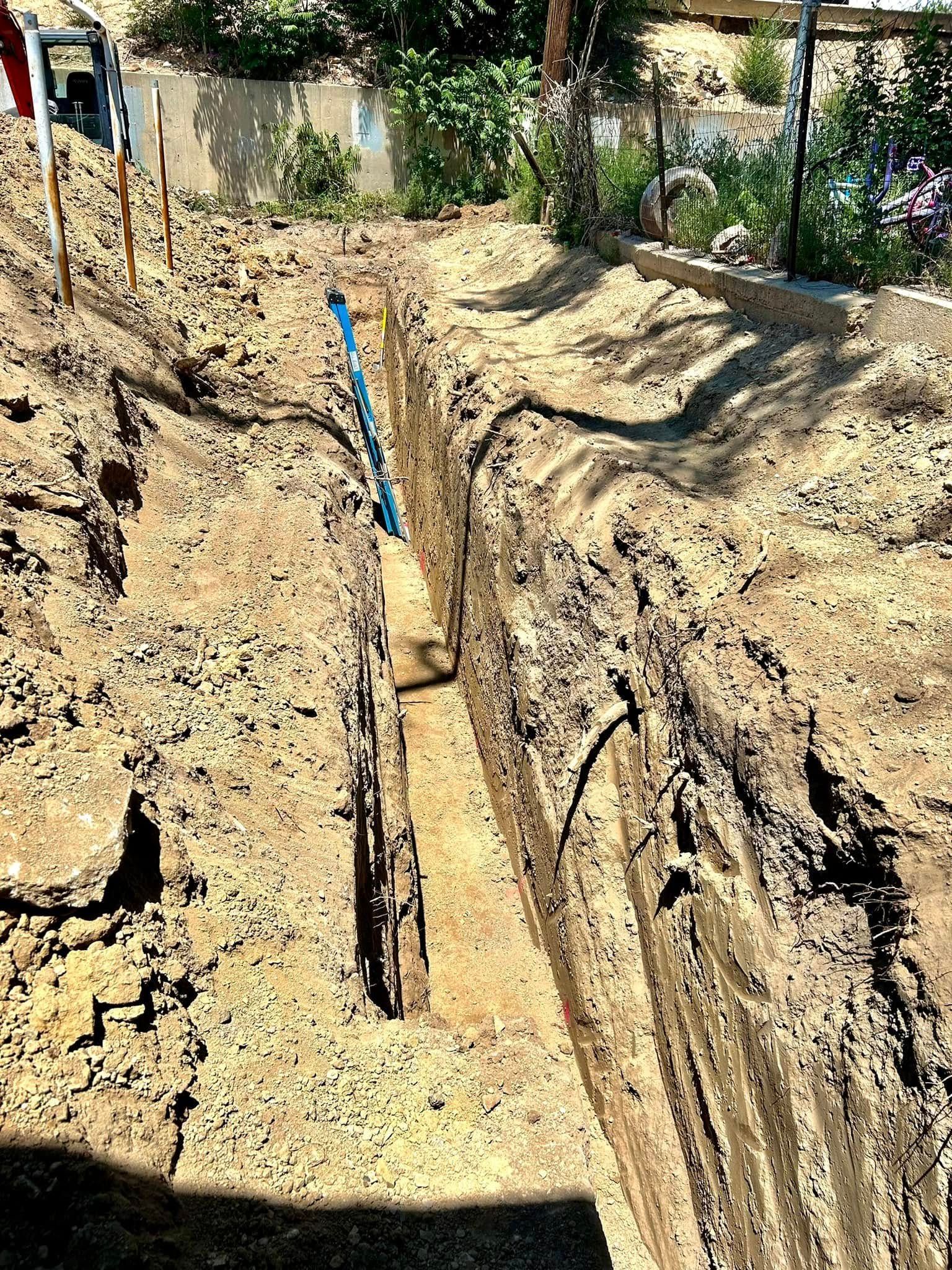 Trench cut into dry soil with a blue pipe, bordered by dirt piles and a garden fence in the background