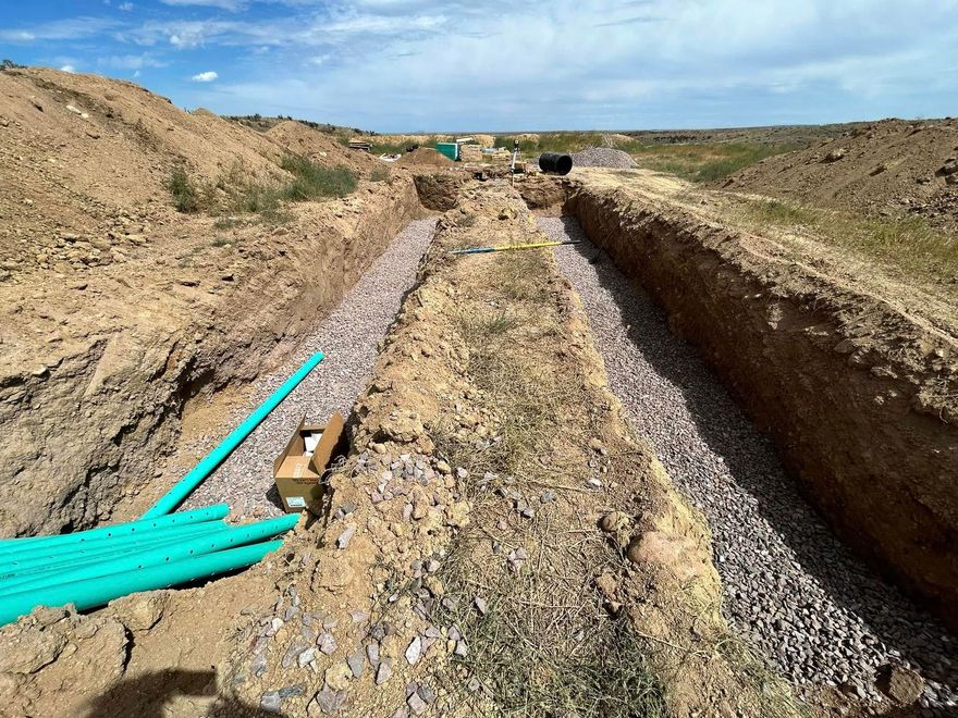 Trenched dirt path with gravel and bright green pipes laid along the left side in a dry construction site