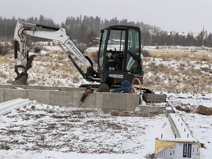 Small excavator working in a snowy field beside a concrete structure and buckets of construction material