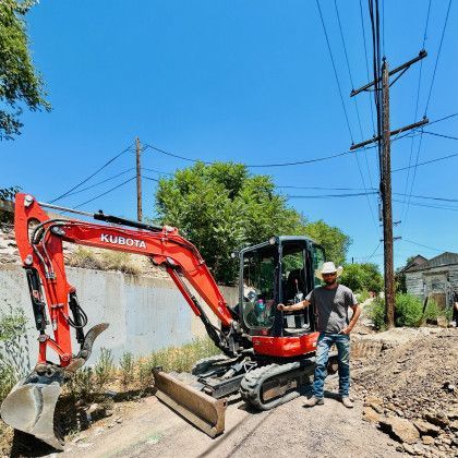 Red Kubota excavator on a dirt road, with a worker standing beside it under a clear blue sky.