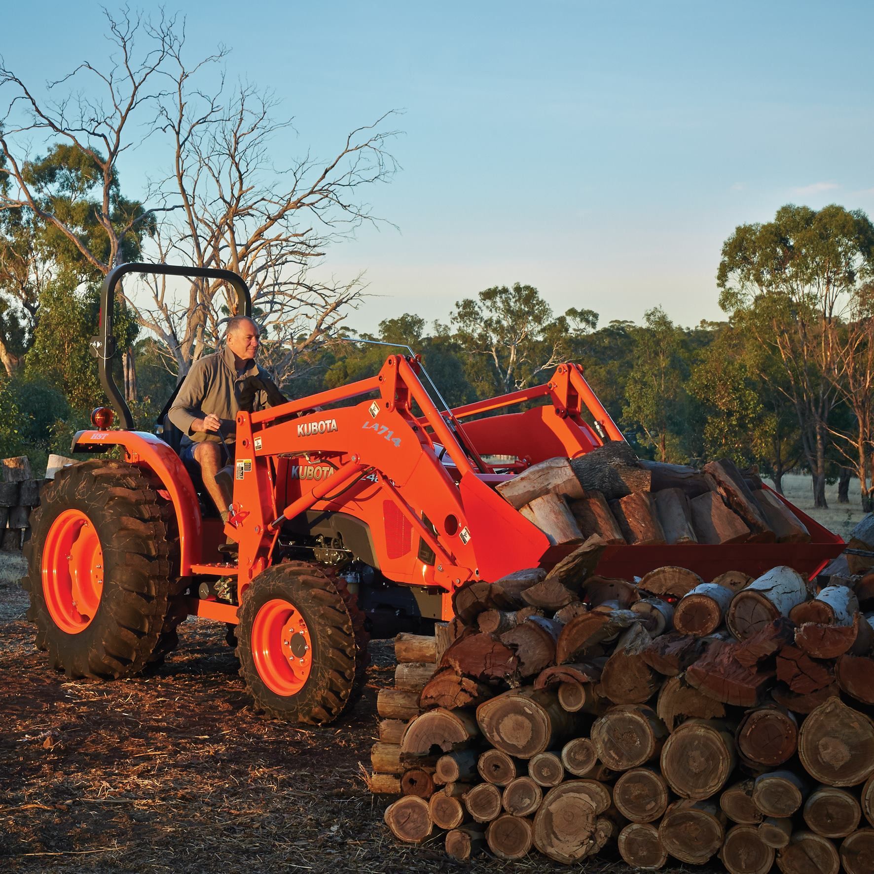 A man is driving an orange tractor next to a pile of logs — Ken Mills Machinery in Kingaroy, QLD