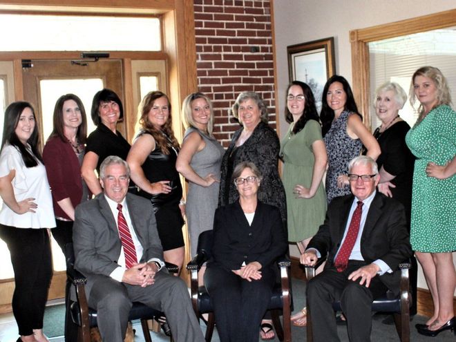 Group of people posing indoors in front of a brick wall, with three seated in the front row.