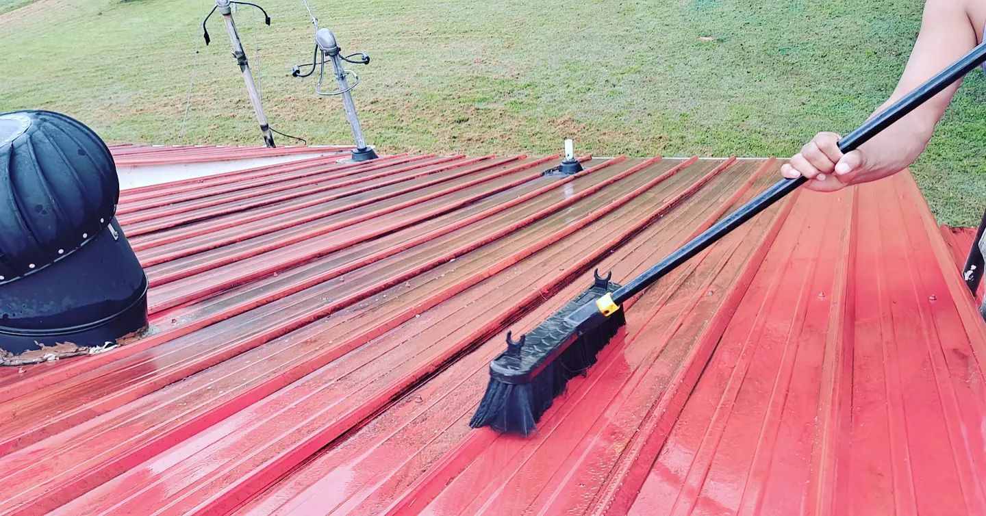 Worker pressure-washing a red metal roof with a black hose and spray wand