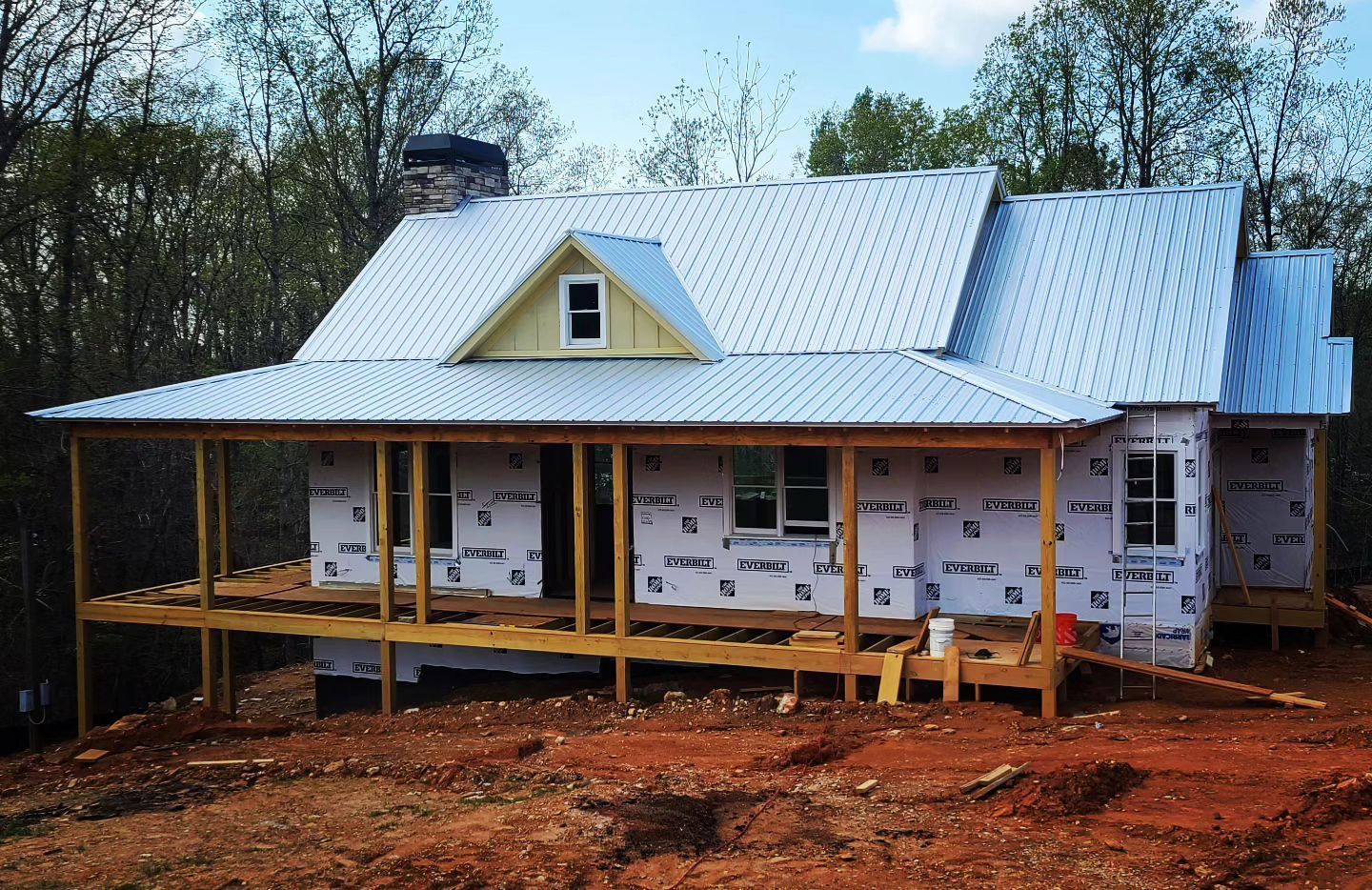 Partially built white house with porch and metal roof on a red dirt lot, surrounded by trees