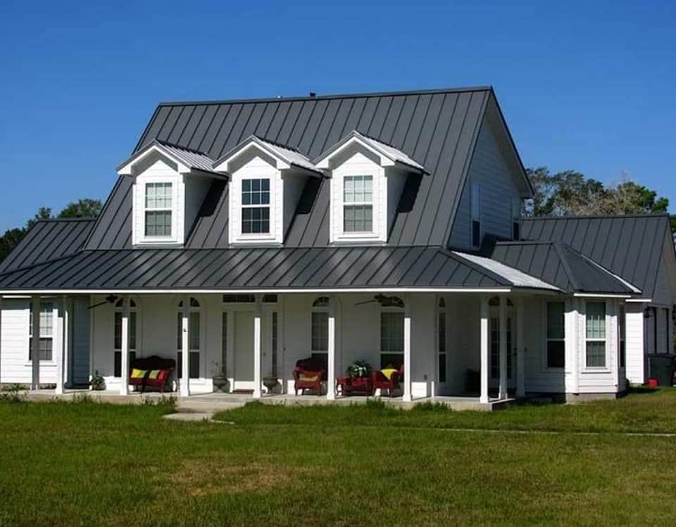 White farmhouse with dark metal roof, front porch, and dormer windows on a grassy lawn.
