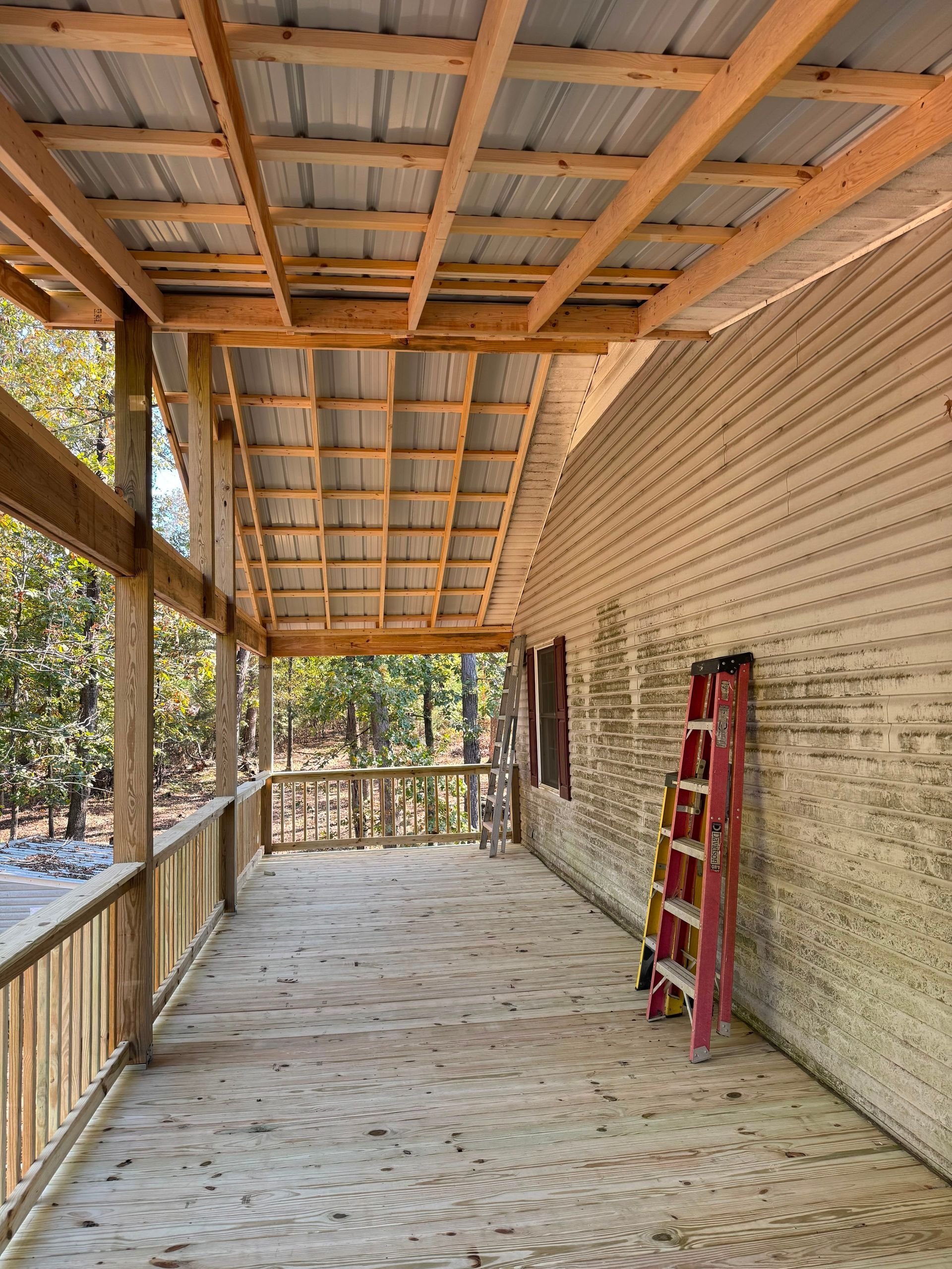 Covered porch under construction with exposed beams, brick wall, and a red ladder leaning against the side.