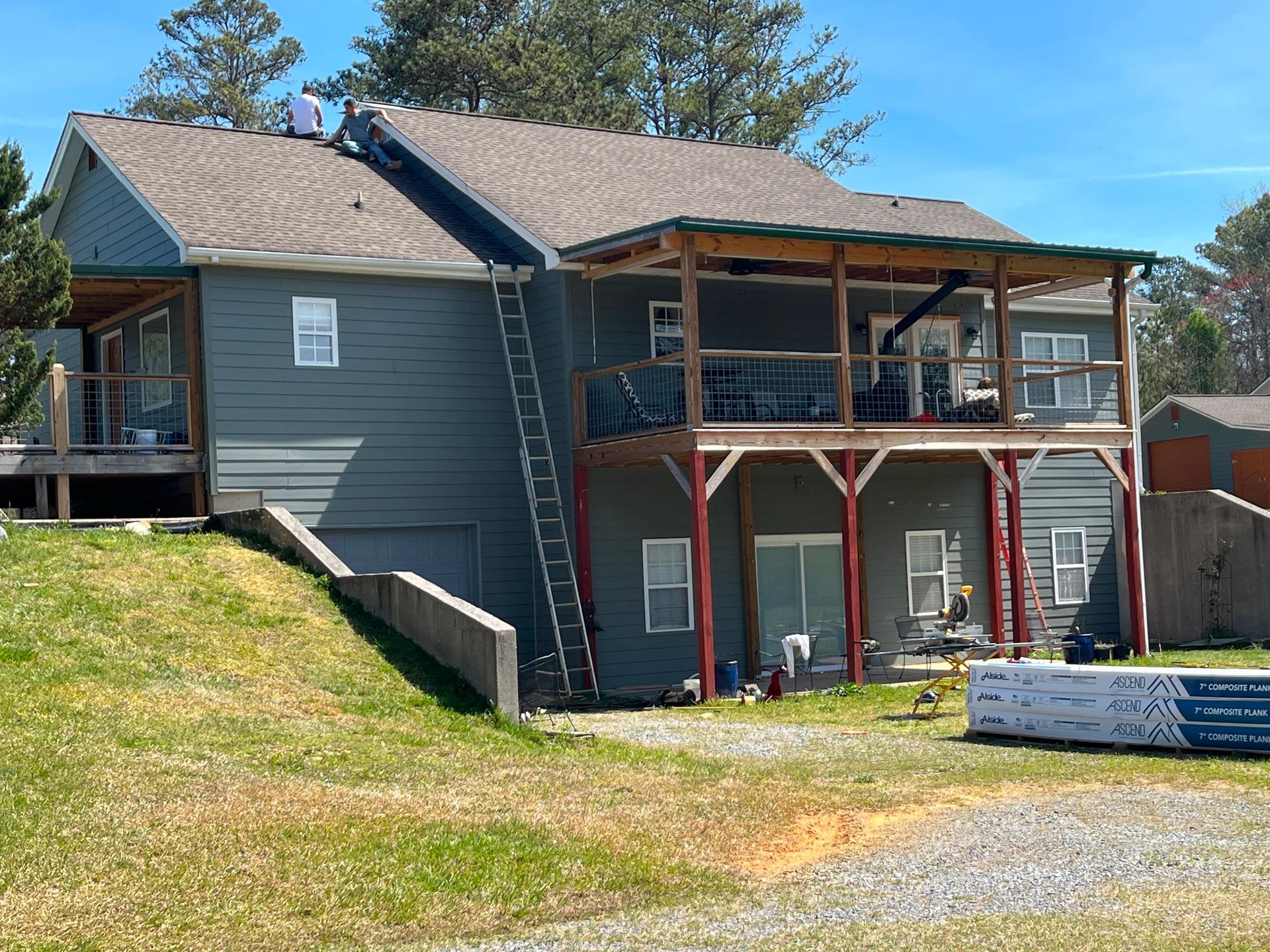 Gray house on a hillside with a covered porch, wooden stairs, and a gravel driveway under trees