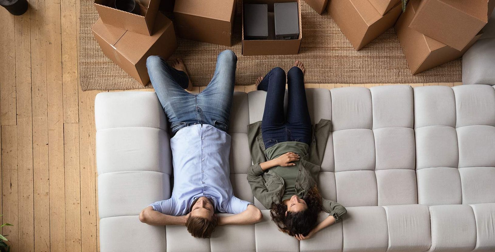 Two people lying on a light gray couch in a living room, viewed from above, surrounded by cardboard boxes.