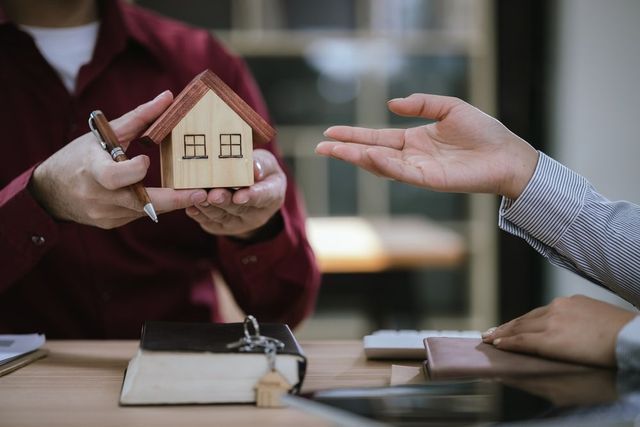 Two people exchange a small house model during a real estate meeting at a table.