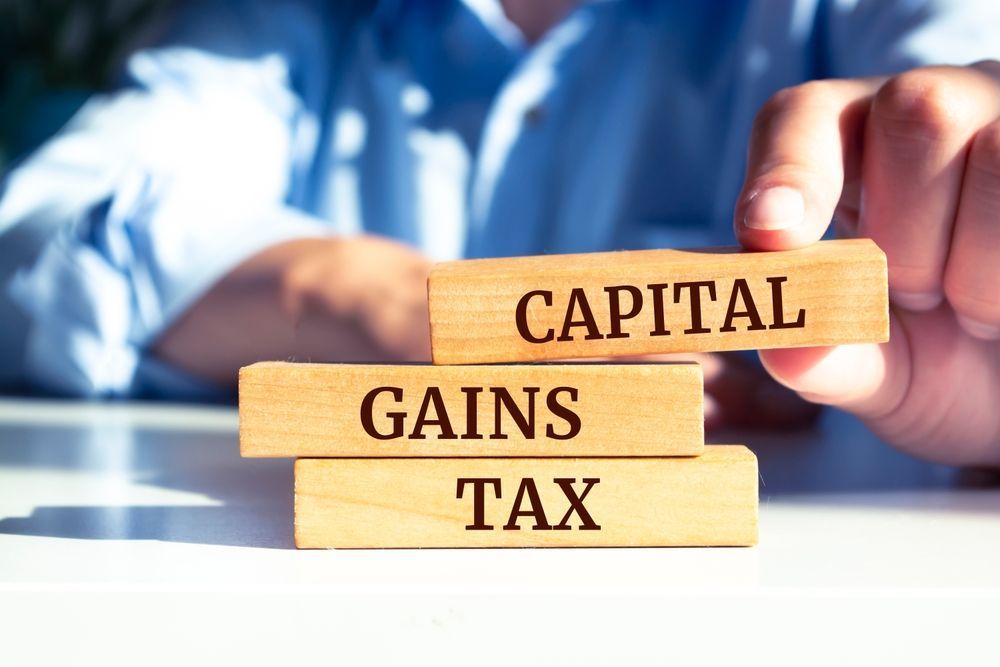 Hands holding wooden blocks labeled “CAPITAL GAINS TAX” on a desk, with a blurred office background.