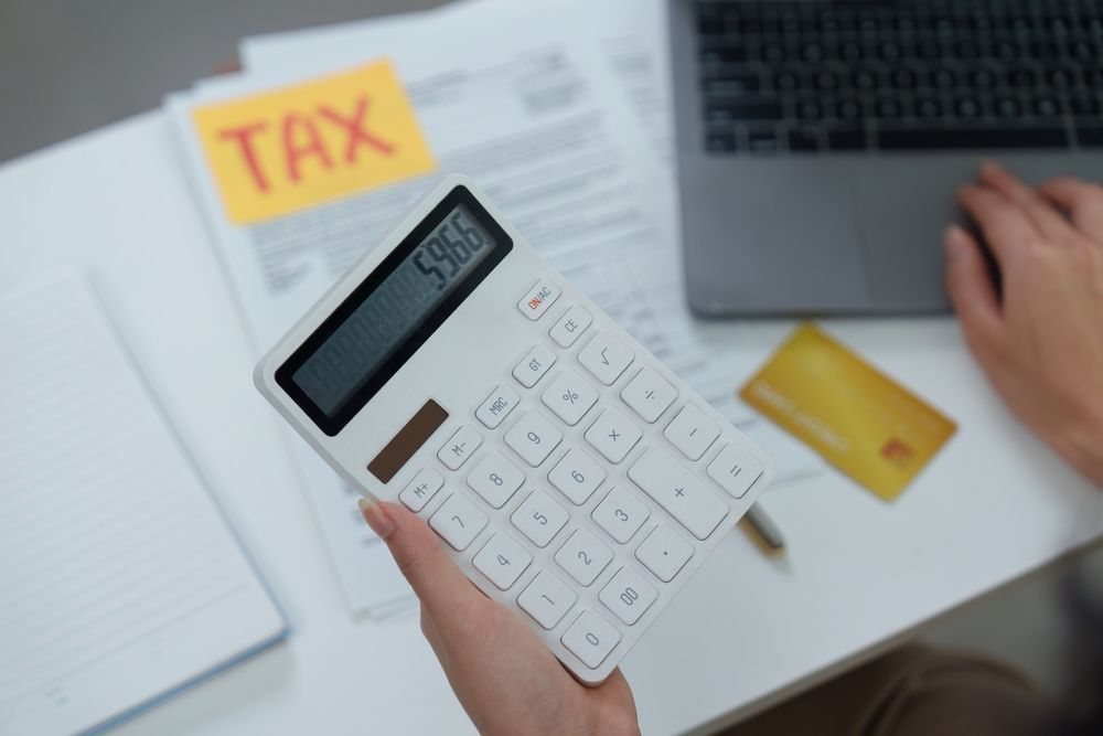 Hand holding calculator beside tax papers, notebook, laptop, and payment card on desk