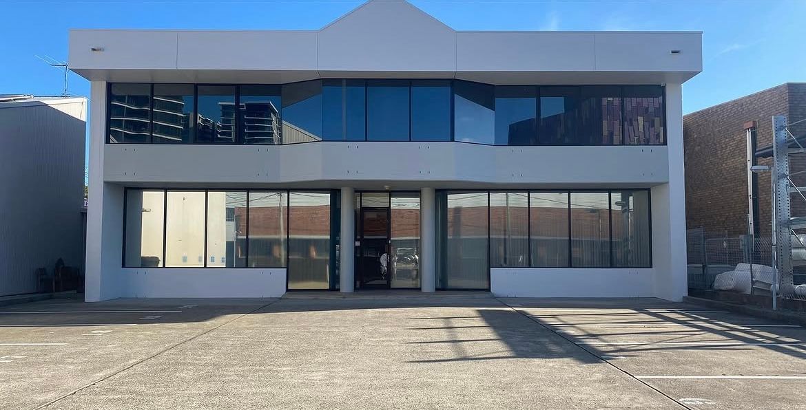 Two-story storefront with large windows and a glass entrance, viewed from a gravel lot in bright sunlight