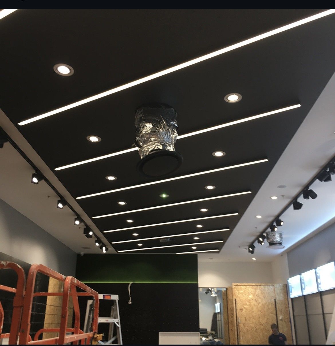 Ceiling under construction with black panels, recessed lights, and ladders in a commercial interior