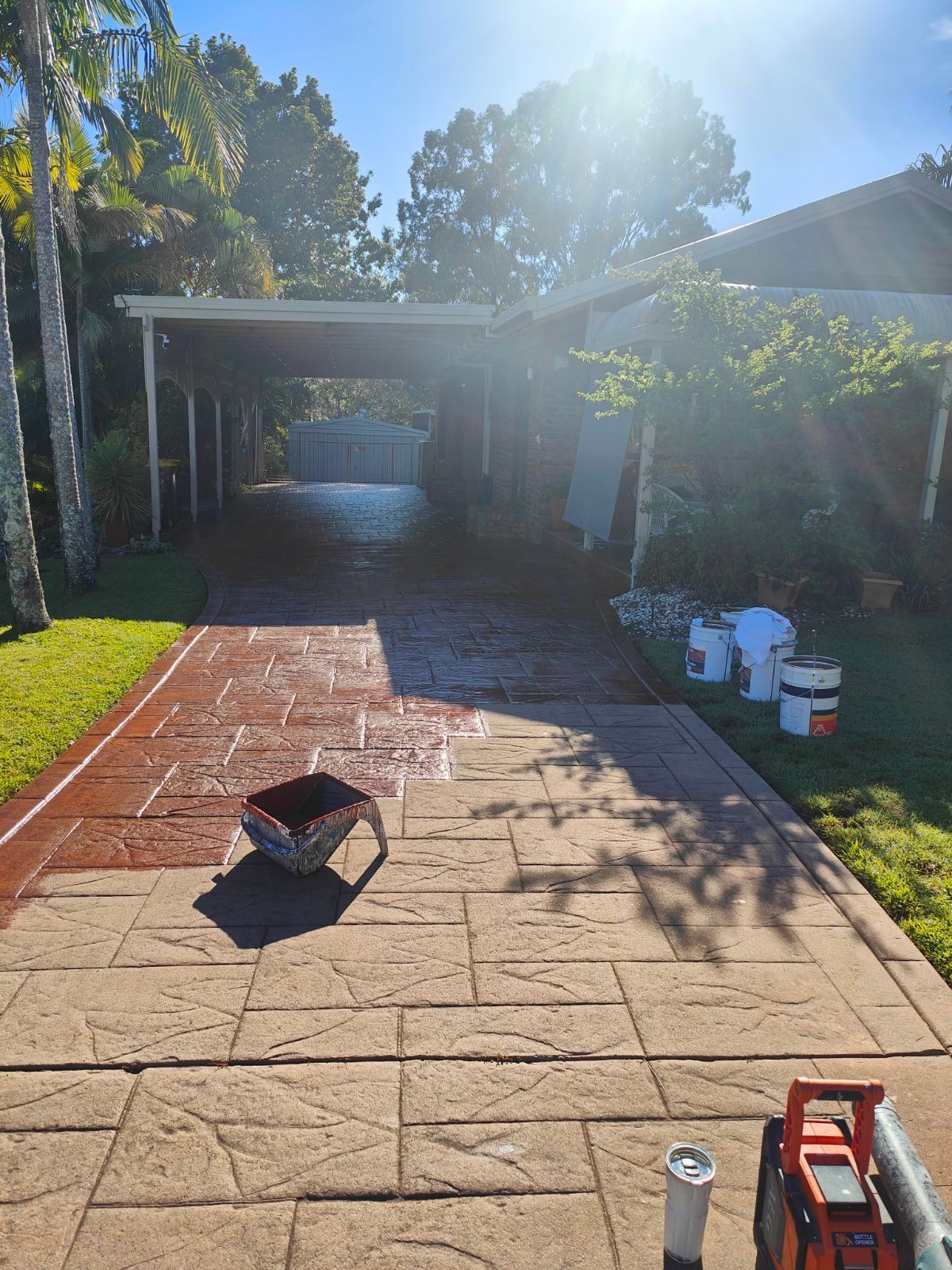 Sunlit backyard patio with paver walkway, tools, buckets, and a carport in the distance