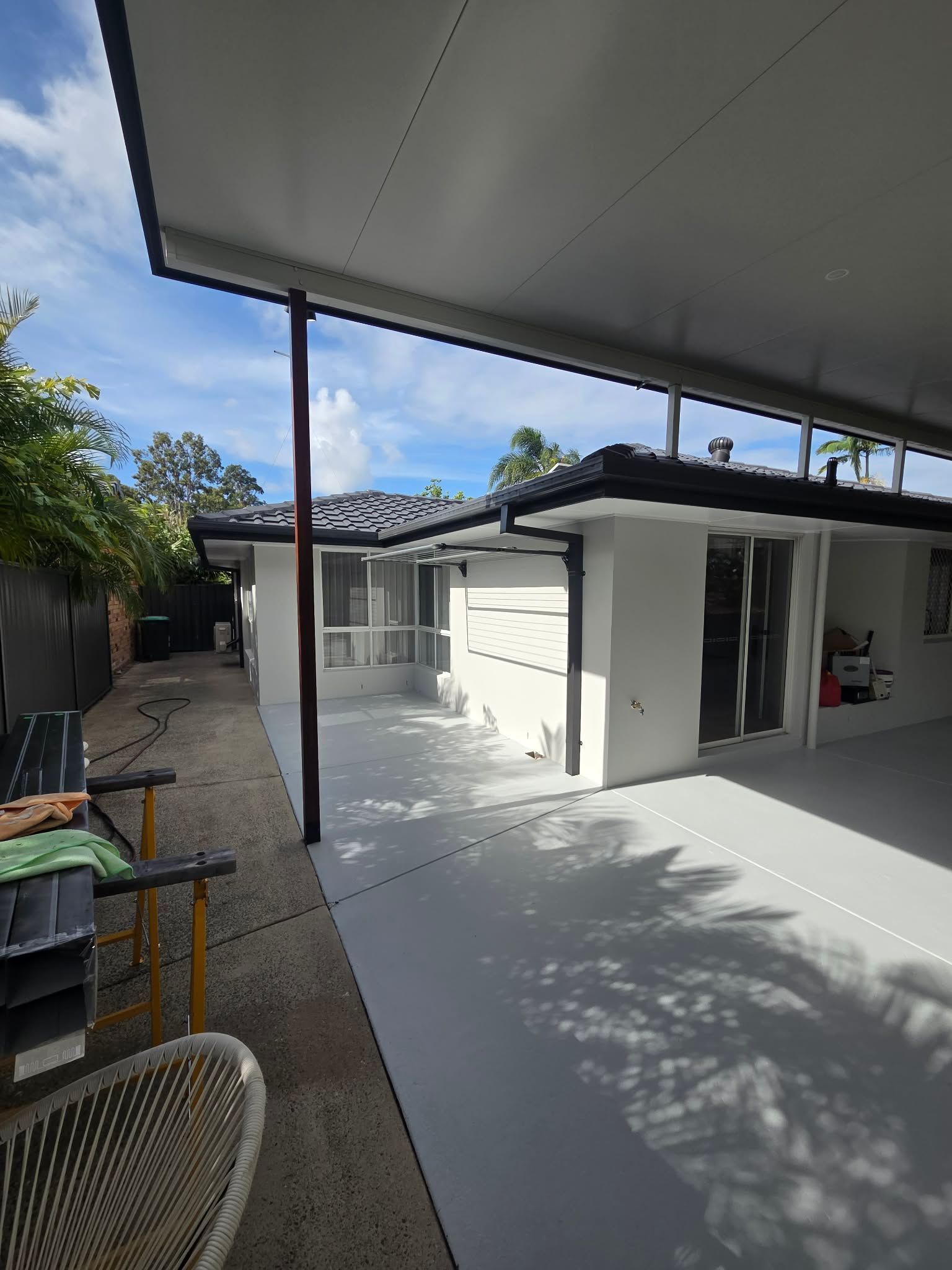 Covered outdoor patio with white flooring, black posts, and a small white utility building in bright sunlight.