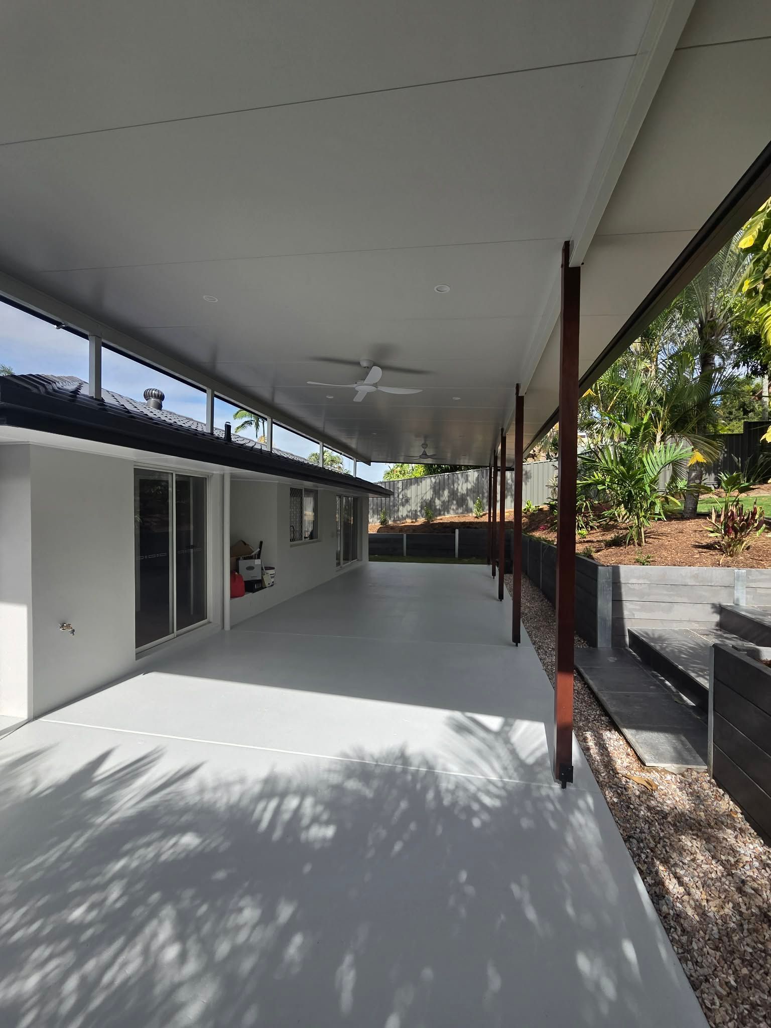 Covered outdoor patio beside a white building, with concrete floor, dark posts, and trees beyond.